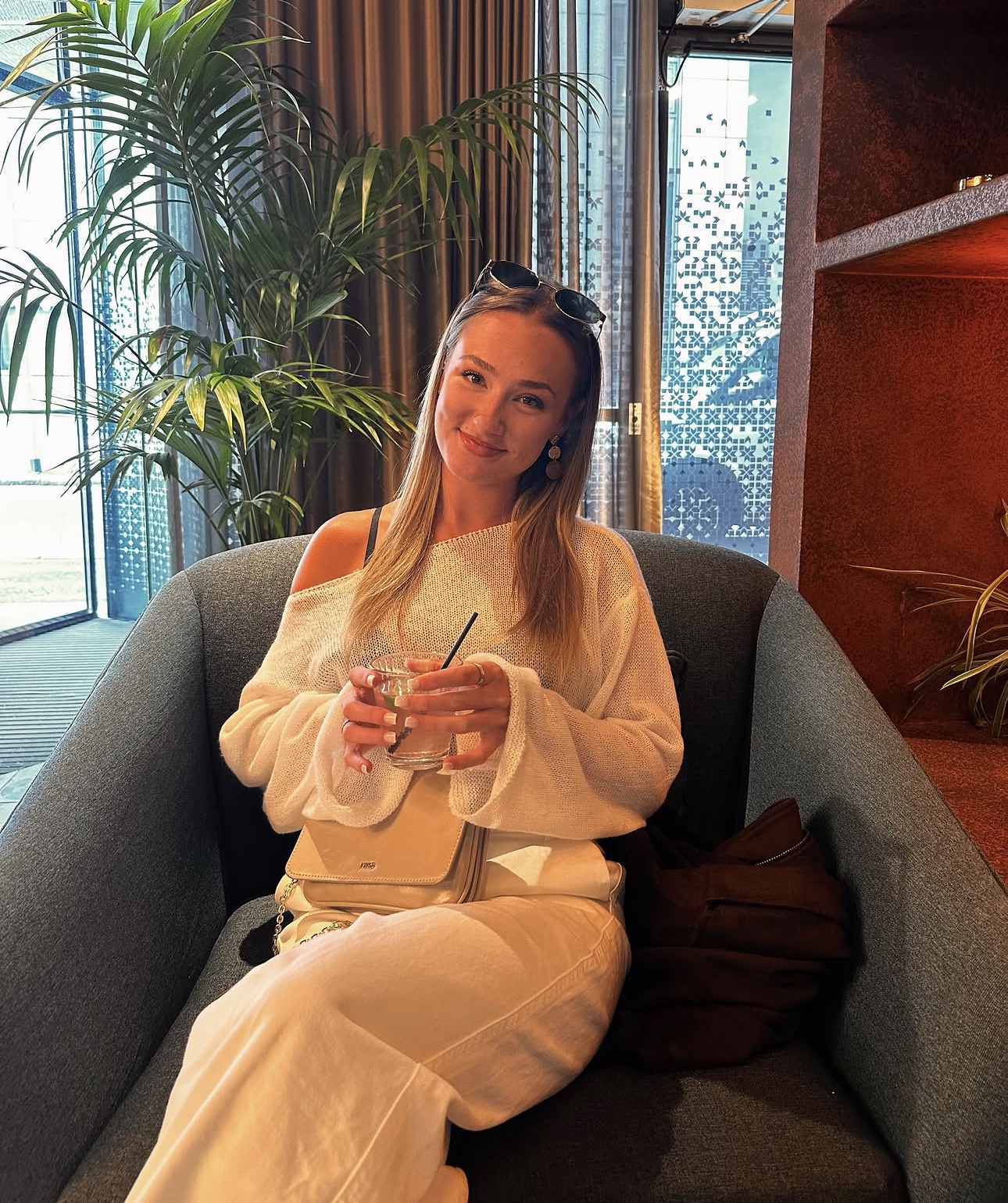 Skye in a cream knit outfit, seated in a lounge chair with a drink